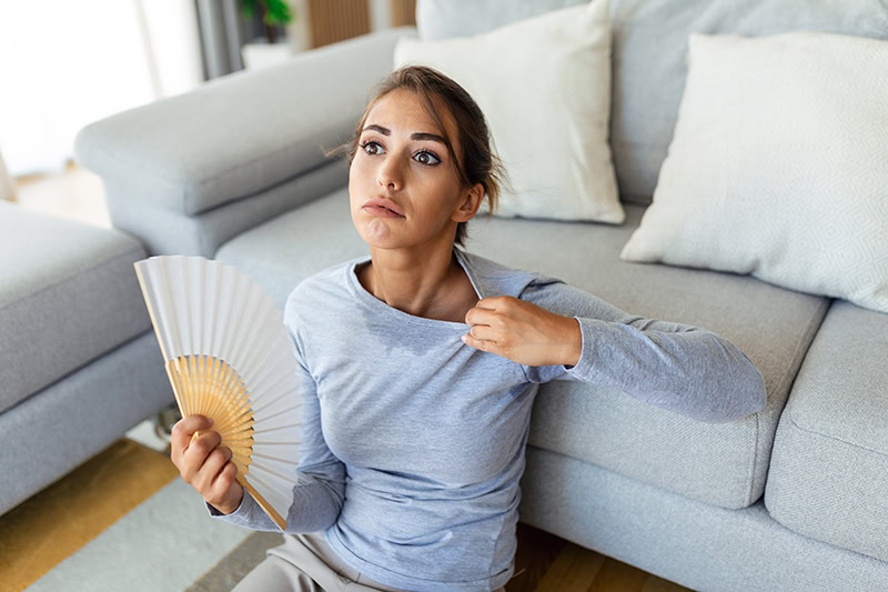 A woman sitting on the floor leaning against a grey sofa, feeling uncomfortably hot and exhausted.