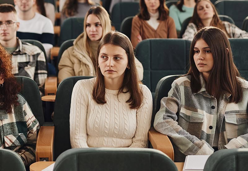 A group of young students sitting in a lecture hall or auditorium, attentively looking forward.