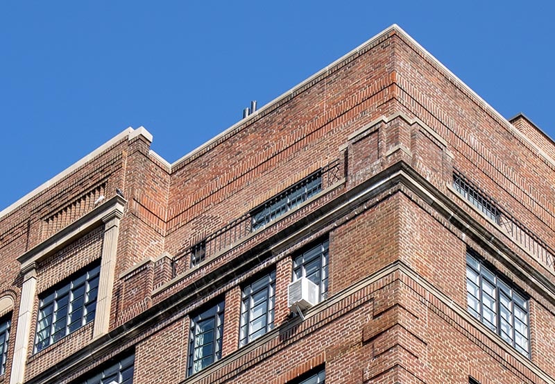 A multi-story red brick façade featuring detailed masonry, decorative stone ledges, rectangular windows, and a single window air conditioning unit mounted in one of the middle openings.